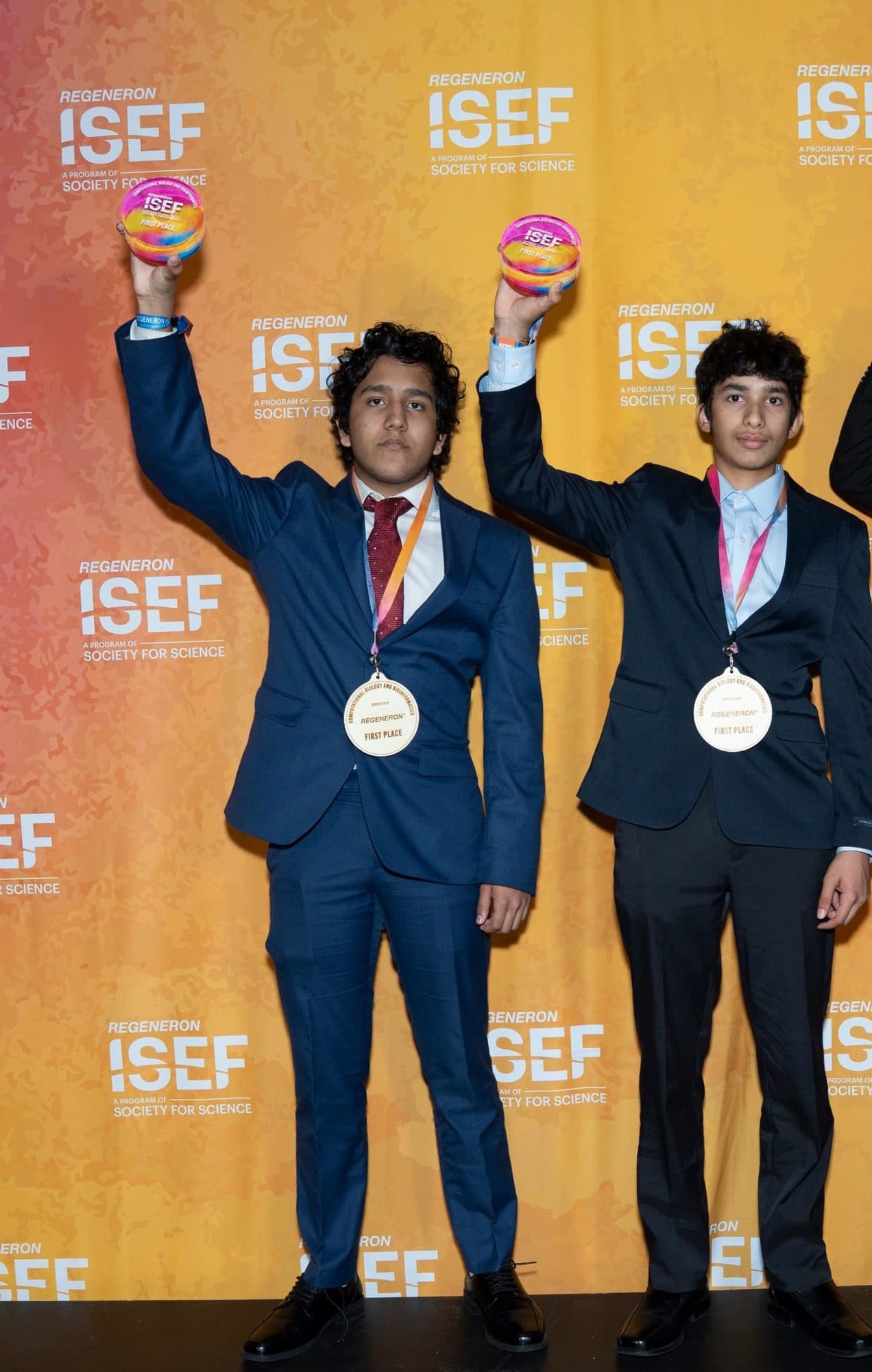 Aryav Das and Samarth Dunakhe raising their Regeneron ISEF trophies, first place medals around their necks, in front of the ISEF backdrop